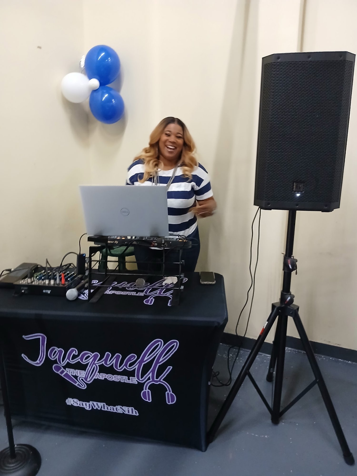 Jacquell The Apostle smiling behind her DJ table with her logo on the table cover