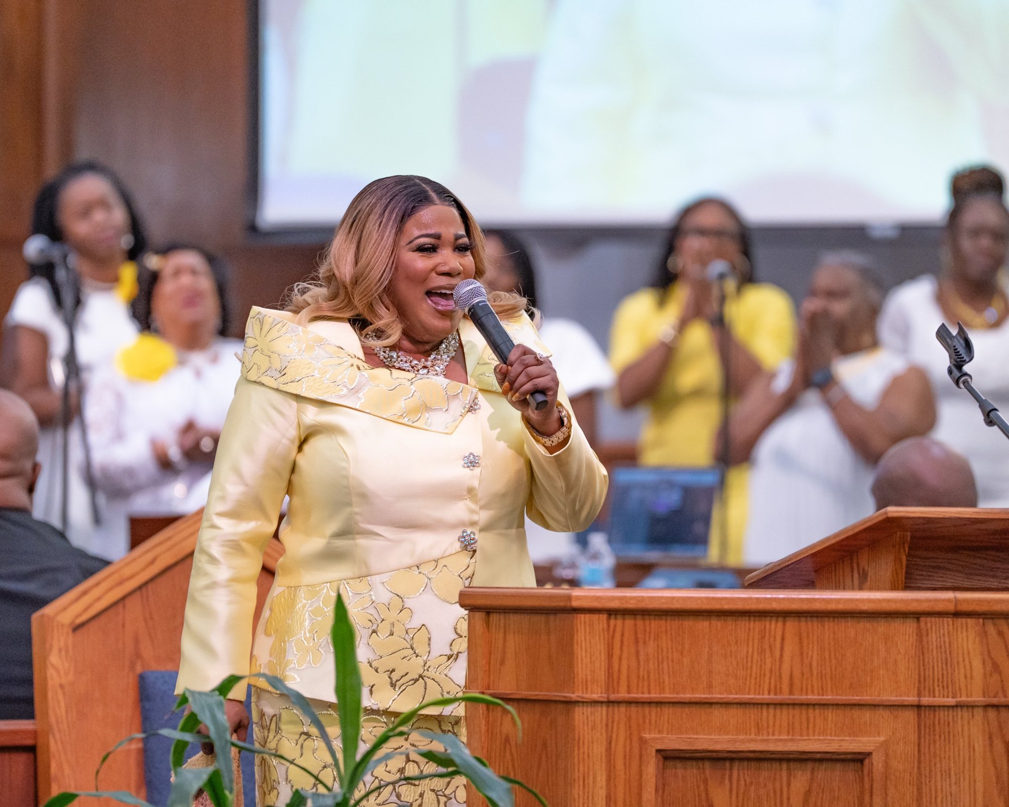 Jacquell The Apostle singing into a microphone at a pulpit with the choir behind her